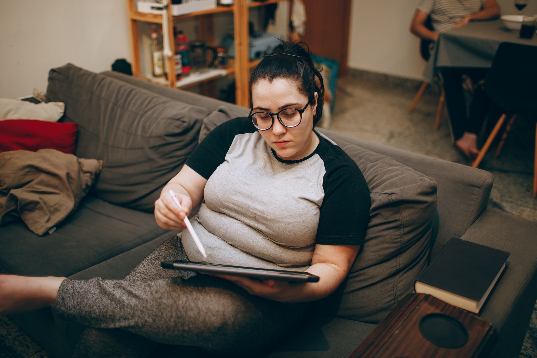 Mujer relajada en un sofá usando una tableta con lápiz digital, en un entorno de trabajo remoto.